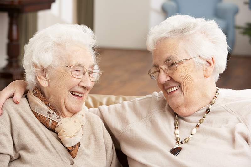 Two senior women friends in a care facility sitting on sofa smiling at each other